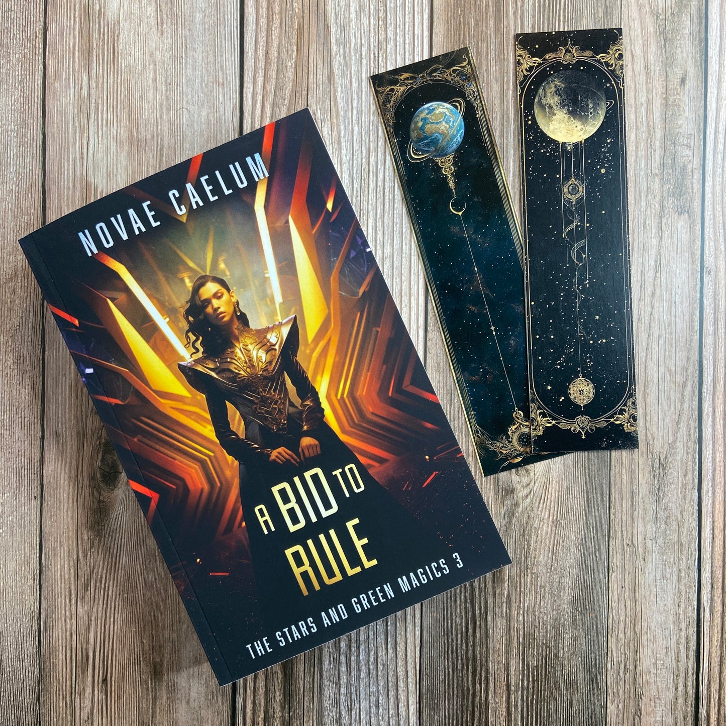 A paperback titled "A Bid to Rule: The Stars and Green Magics - Book 3" by Novae Caelum, featuring a cover image of a nonbinary individual in a futuristic gown standing with glowing waves in the background; there are also 2 series-related bookmarks arranged beside the book.