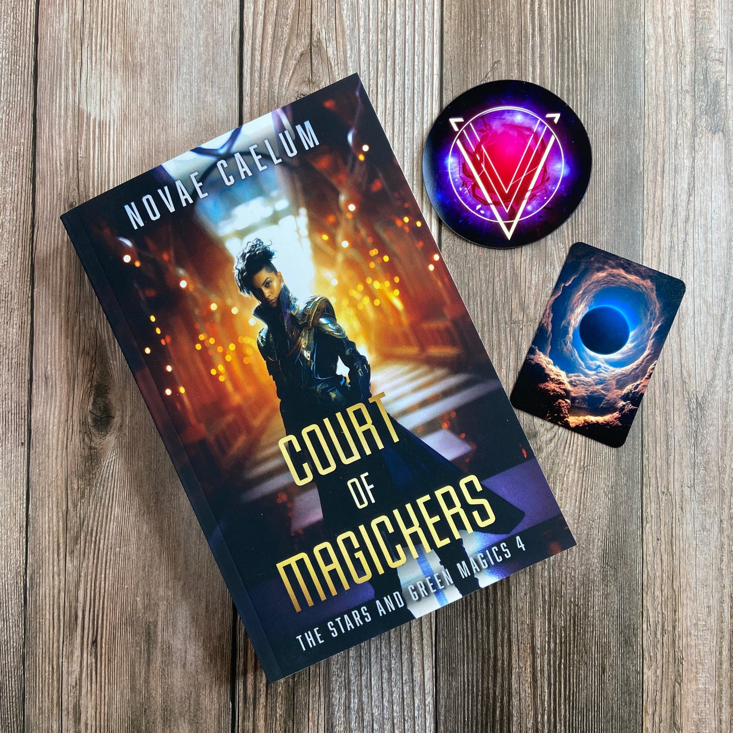 A paperback titled "A Bid to Rule: The Stars and Green Magics - Book 3" by Novae Caelum, featuring a cover image of a nonbinary individual in a futuristic gown standing with glowing waves in the background; there are also 2 series-related cards arranged beside the book.
