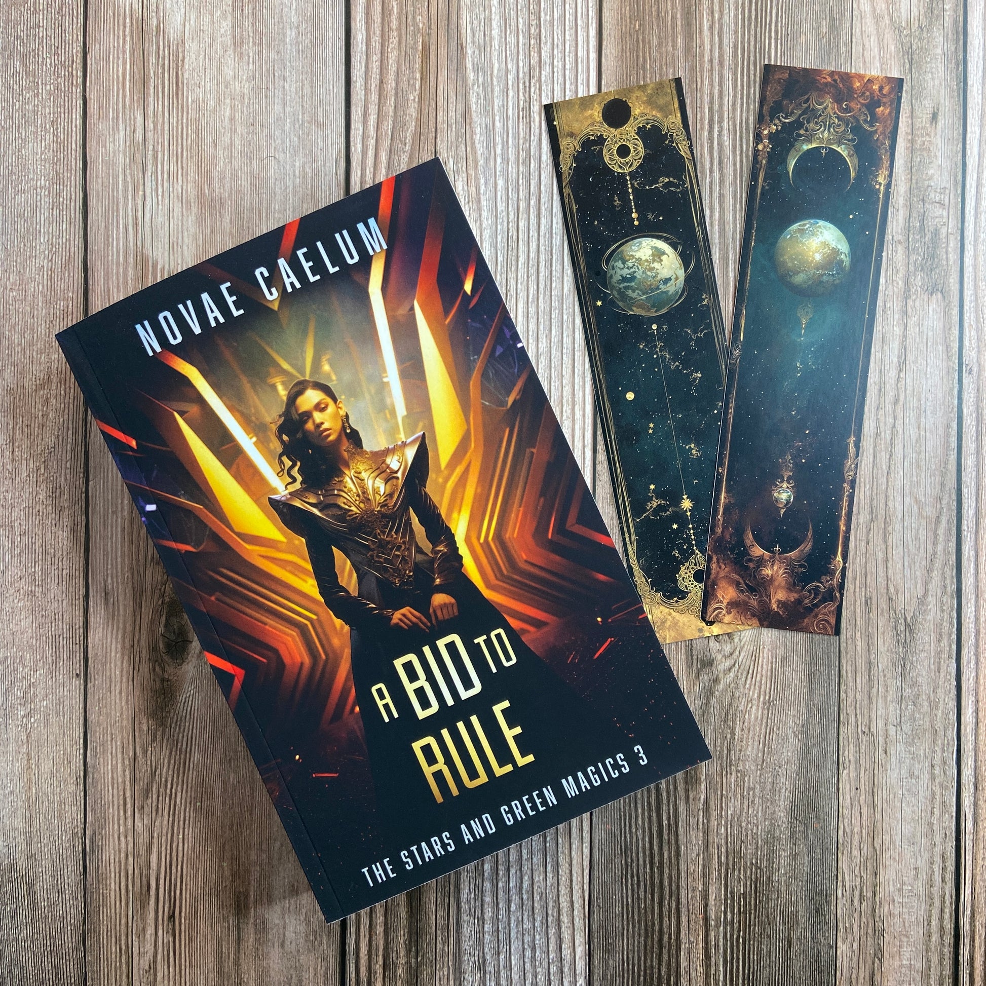 A paperback titled "A Bid to Rule: The Stars and Green Magics - Book 3" by Novae Caelum, featuring a cover image of a nonbinary individual in a futuristic gown standing with glowing waves in the background; there are also 2 series-related bookmarks arranged beside the book.