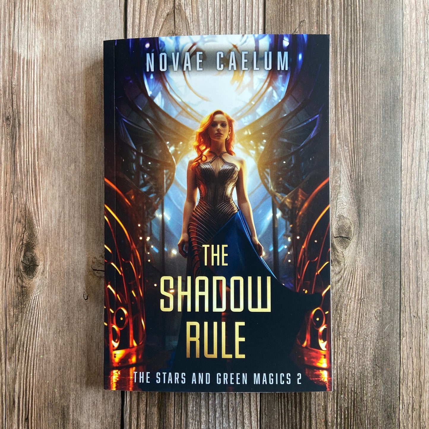 A paperback titled "The Shadow Rule: The Stars and Green Magics - Book 2" by Novae Caelum, featuring a cover image of a woman in a sleeveless, futuristic gown standing with illuminated, circular windows in the background.