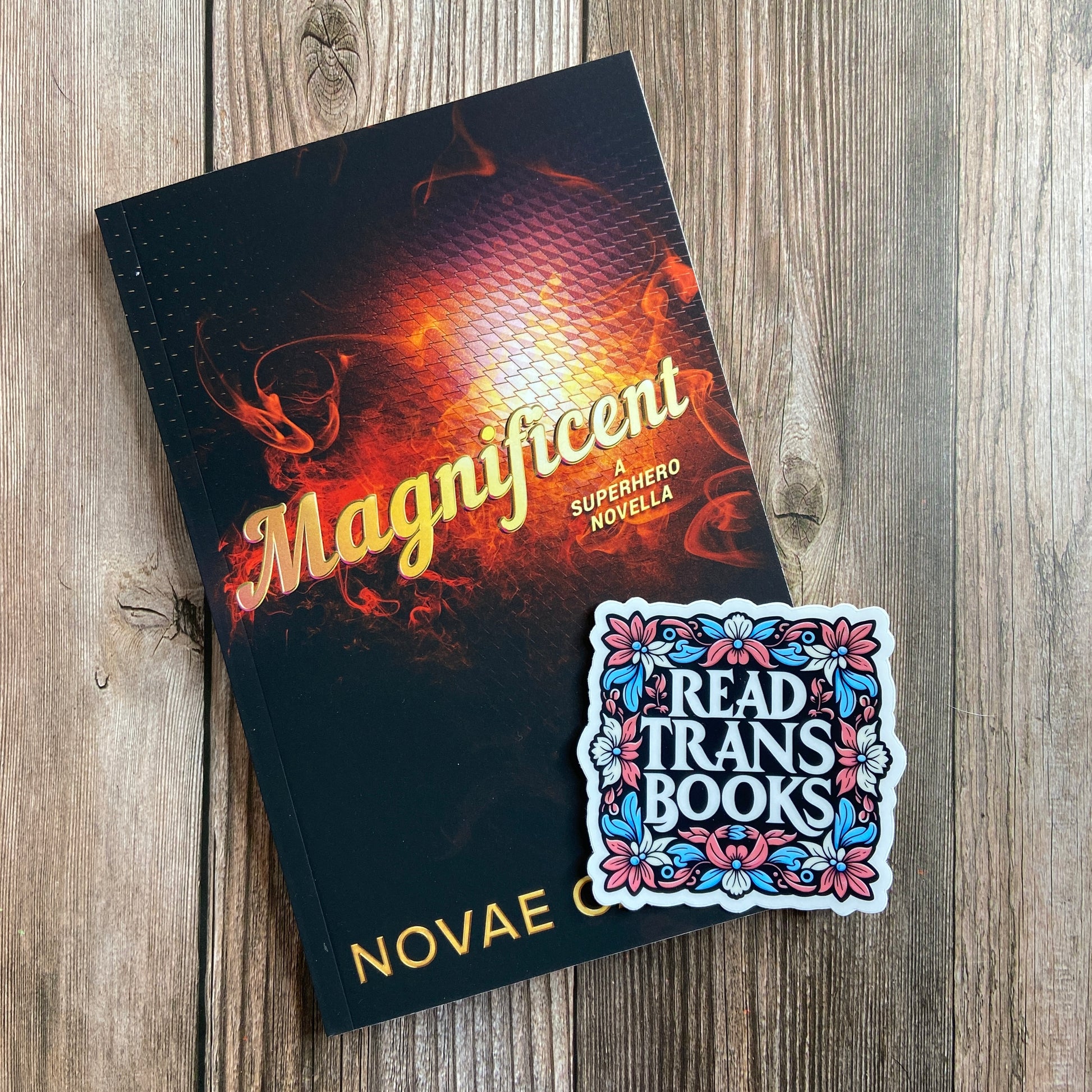 A paperback titled "Magnificient: A Nonbinary Superhero Novella" by Novae Caelum with an image of swirling red smoke on a black backdrop; there is also a series-related sticker arranged atop the book.