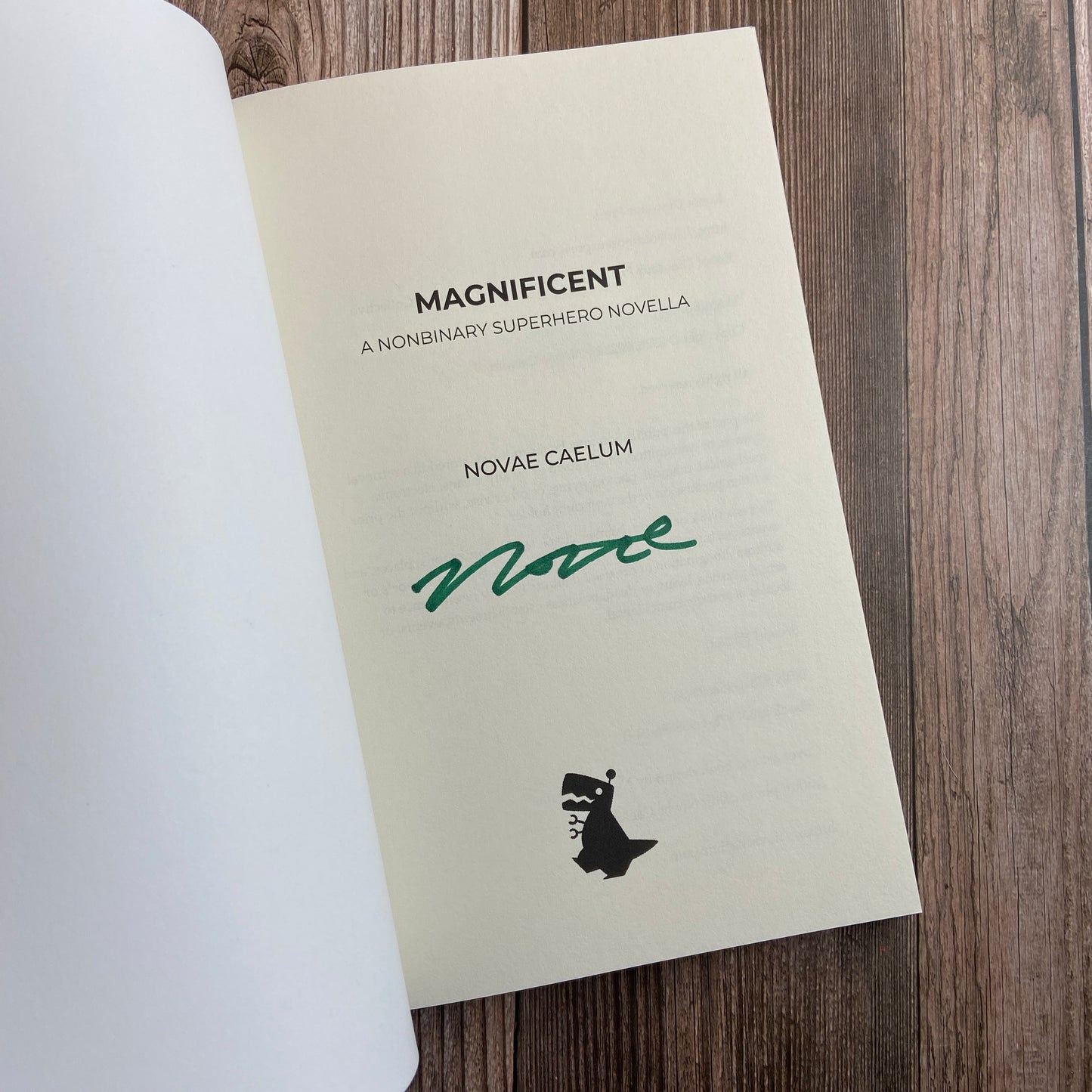 A paperback (titled "Magnificient: A Nonbinary Superhero Novella" by Novae Caelum) opened to display the author-signed title page.