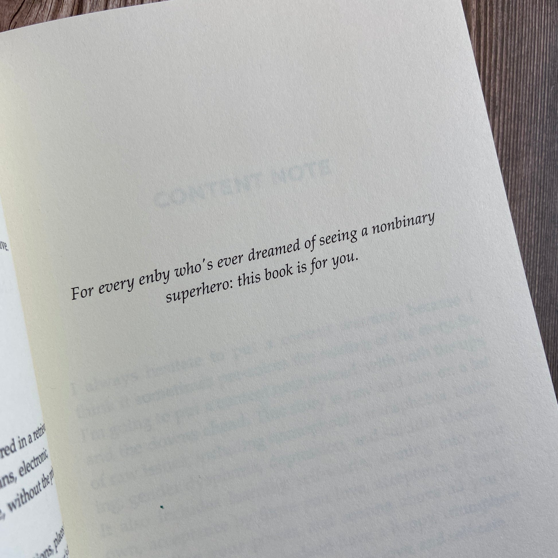 A paperback (titled "Magnificient: A Nonbinary Superhero Novella" by Novae Caelum) opened to display the dedication page, which reads: "For every enby who's ever dreamed of seeing a nonbinary superhero: this book is for you."
