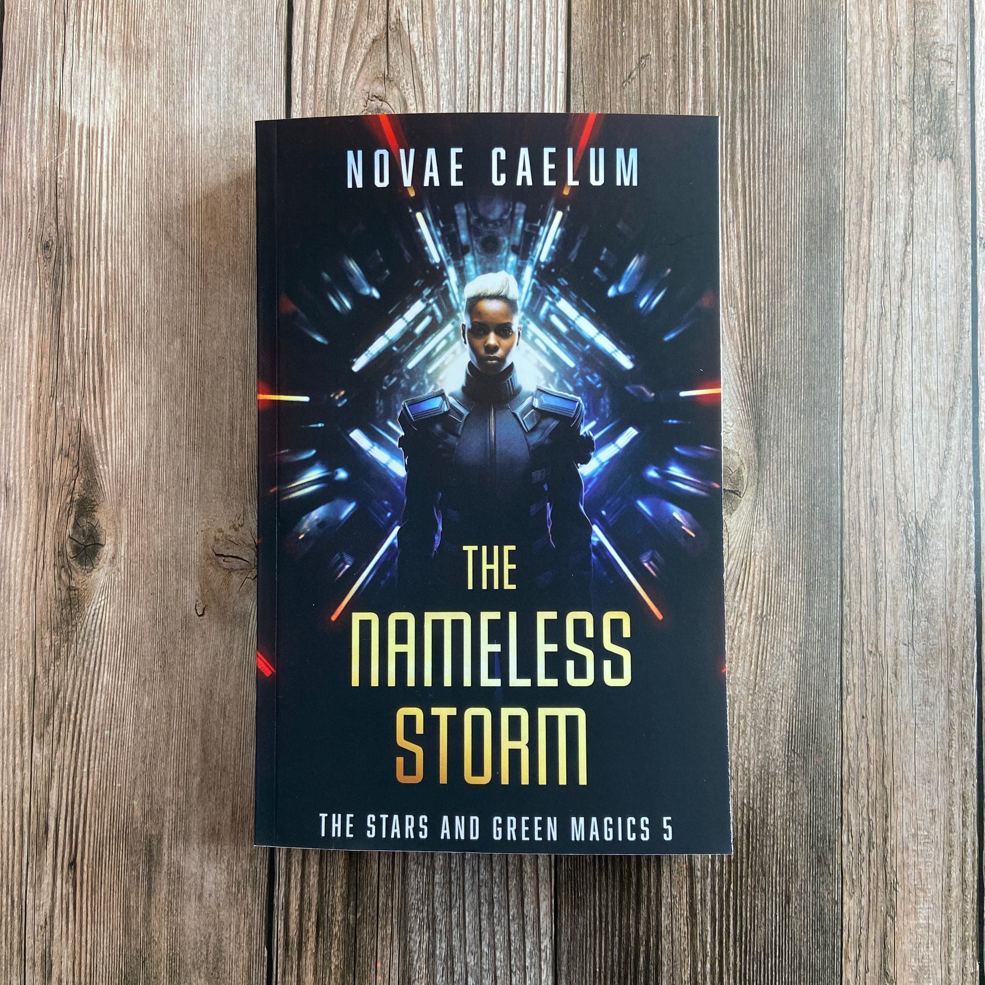 A hardcover book titled "SIGNED The Nameless Storm: The Stars and Green Magics Book 5" from Novae Caelum, featuring a royal sibling in a futuristic outfit, displA paperback titled "The Nameless Storm: The Stars and Green Magics -  Book 5" by Novae Caelum, featuring a cover image of a nonbinary individual in futuristic armor standing before a tunnel of glowing lights in the background.ayed on a wooden surface.
