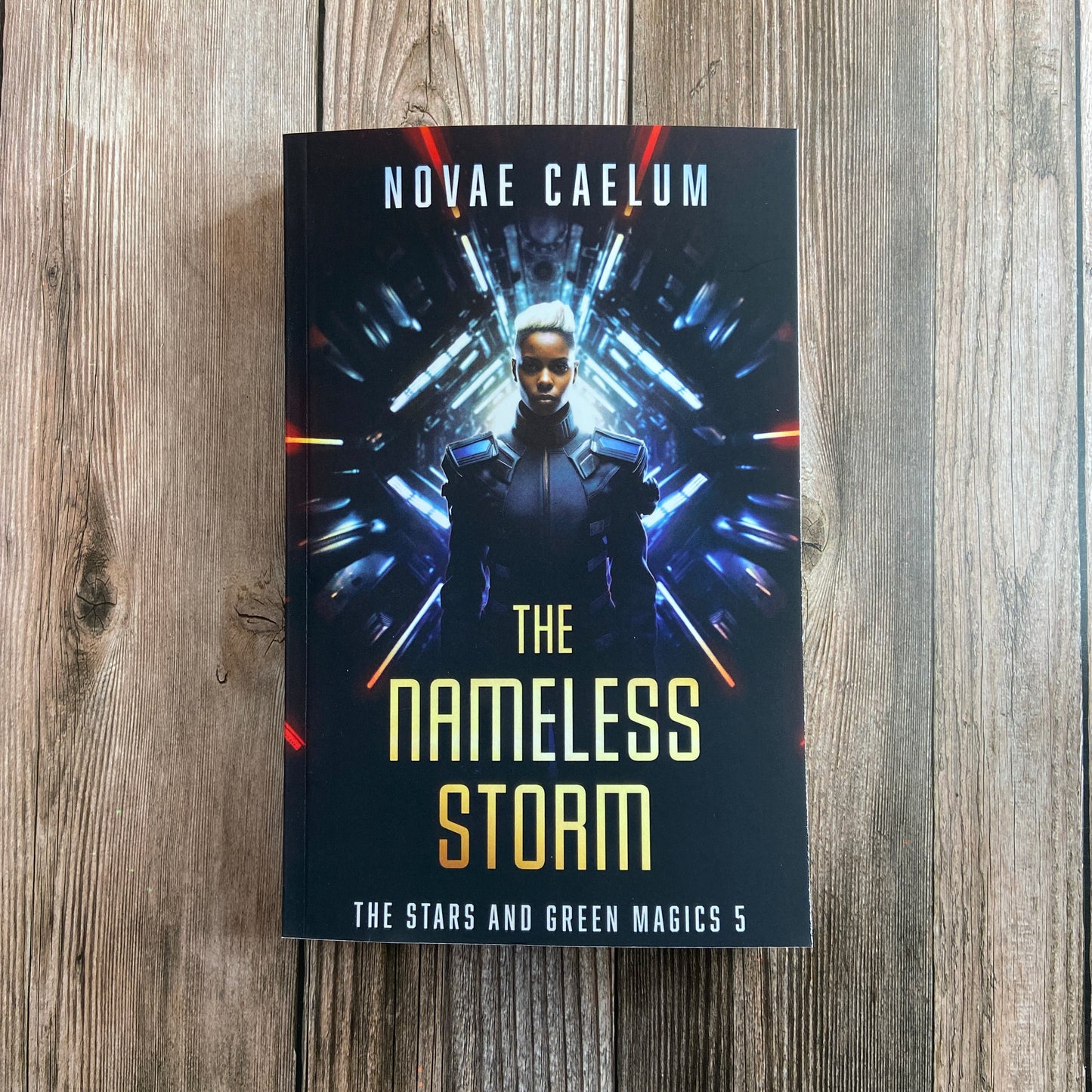 A hardcover book titled "SIGNED The Nameless Storm: The Stars and Green Magics Book 5" from Novae Caelum, featuring a royal sibling in a futuristic outfit, displA paperback titled "The Nameless Storm: The Stars and Green Magics - Book 5" by Novae Caelum, featuring a cover image of a nonbinary individual in futuristic armor standing before a tunnel of glowing lights in the background.ayed on a wooden surface.