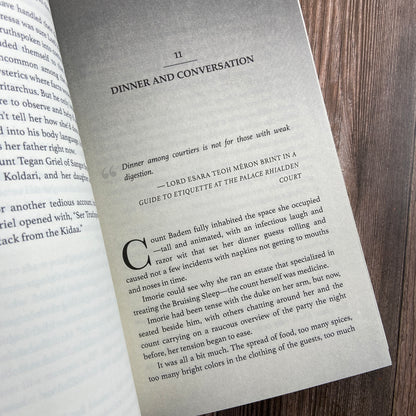 A paperback (titled "The Shadow Rule: The Stars and Green Magics - Book 2" by Novae Caelum) opened to display the first page of "Chapter 11: Dinner and Conversation".