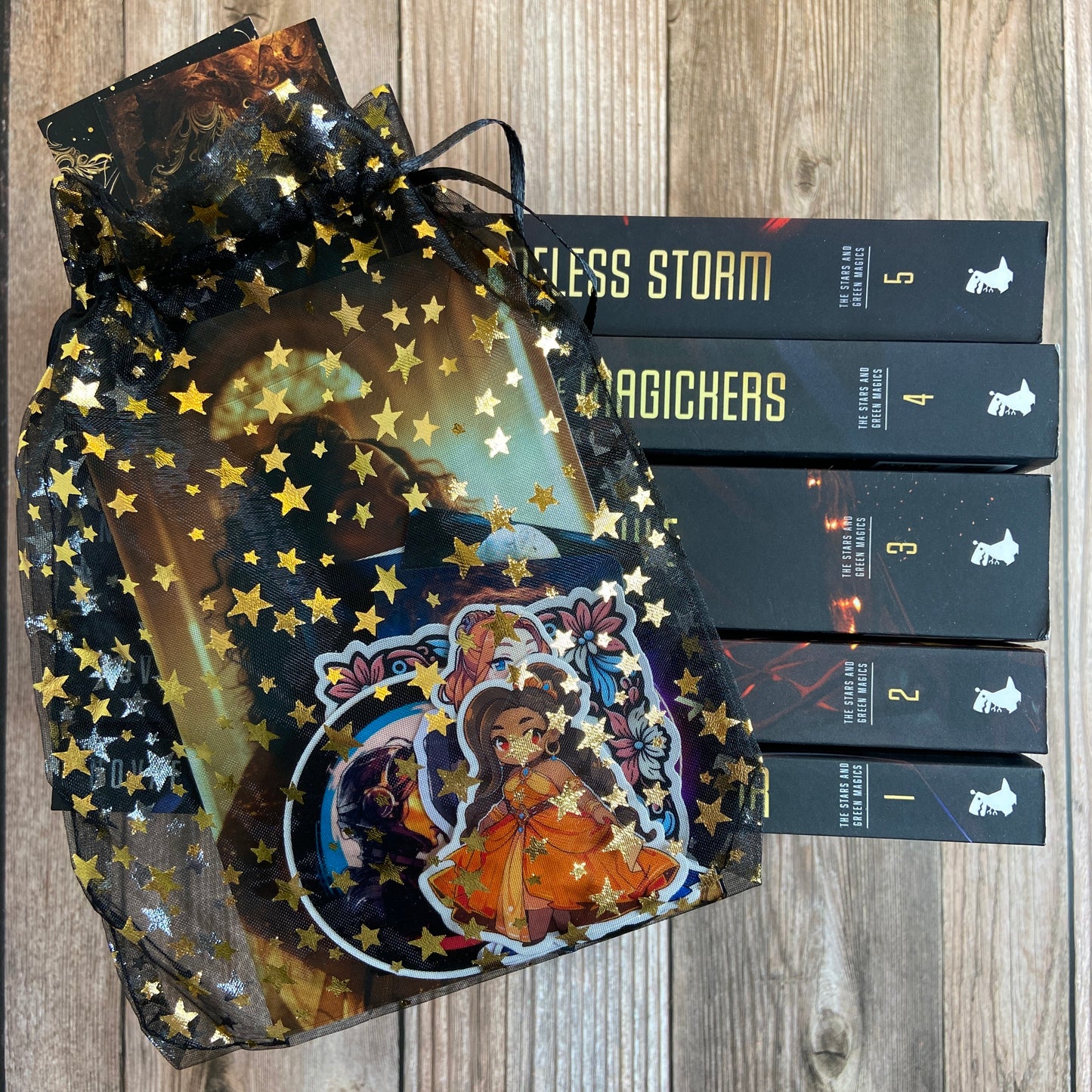 A star-patterned gift bag with an anime-style character sticker next to a Novae Caelum SIGNED Paperback Deluxe Swag Bundle: The Stars and Green Magics (Books 1-5) set of books, renowned for their sapphic arranged marriage plot, rests on a wooden surface.