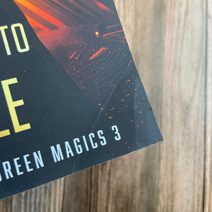 Corner of a Novae Caelum book titled "To Light" with a subtitle "Green Magics 3: Space Magic," set against a wooden background.