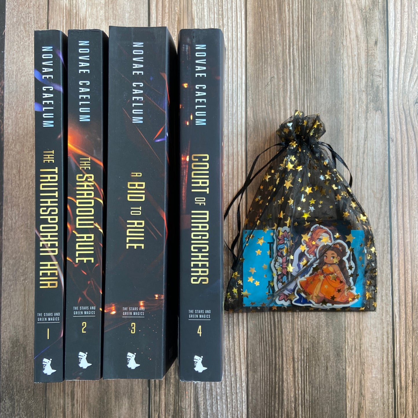 Bundle of 4 paperbacks (Books 1-4) from "The Stars and Green Magics" series by Novae Caelum displaying the spines, along with a black, starry mesh gift bag filled with a variety of series-related stickers. This bundle is part of the "SCRATCH AND DENT - SIGNED" collection.
